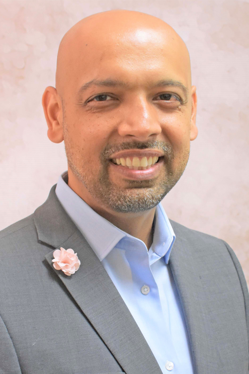 Man wearing a grey suit and light blue shirt smiles at camera.